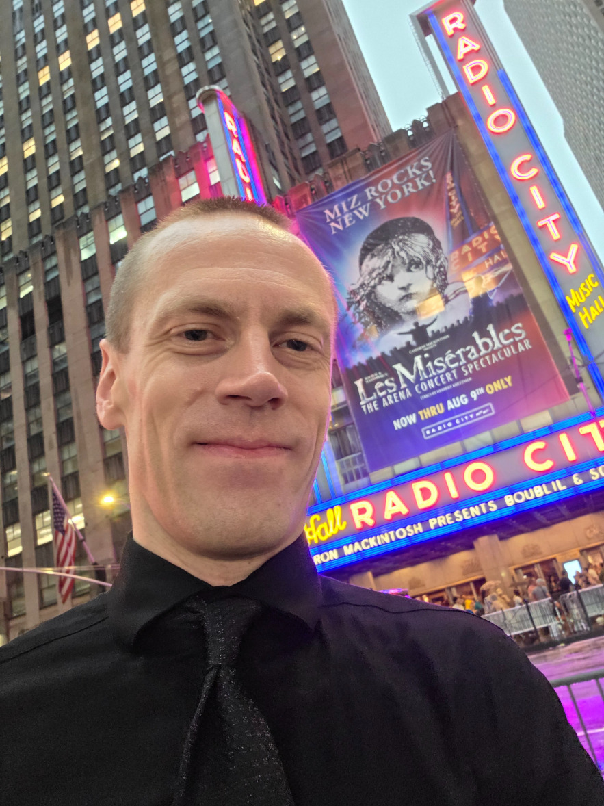 A photo of me in a black dress shirt and a black tie, with a pattern on the tie, standing in front of Radio City Music Hall in New York City in August of 2026.